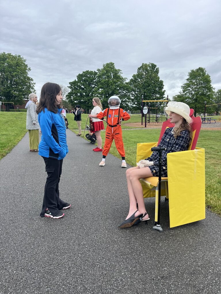 Student speaks to another student dressed as Queen Elizabeth II on a throne. 