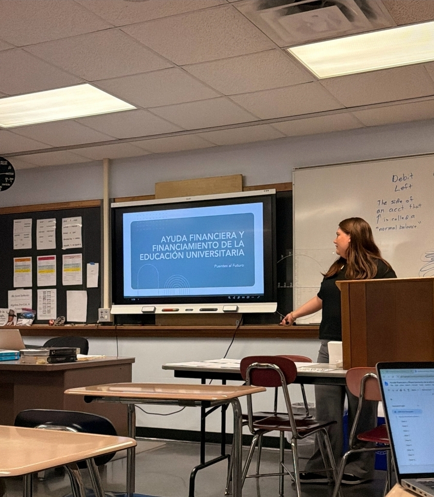 Woman at podium in classroom with presentation on screen. Students seated with computers at tables.
