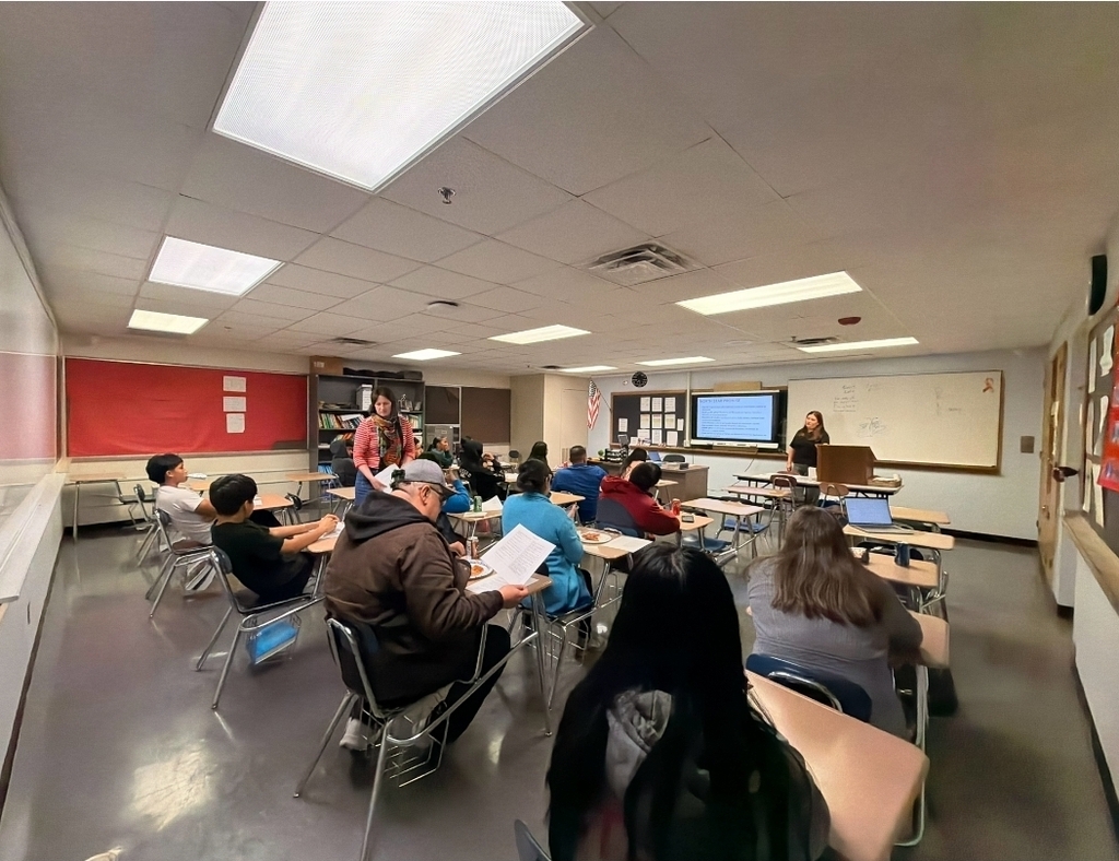 Classroom with seated students and teacher standing at front of room. Some students use laptops.