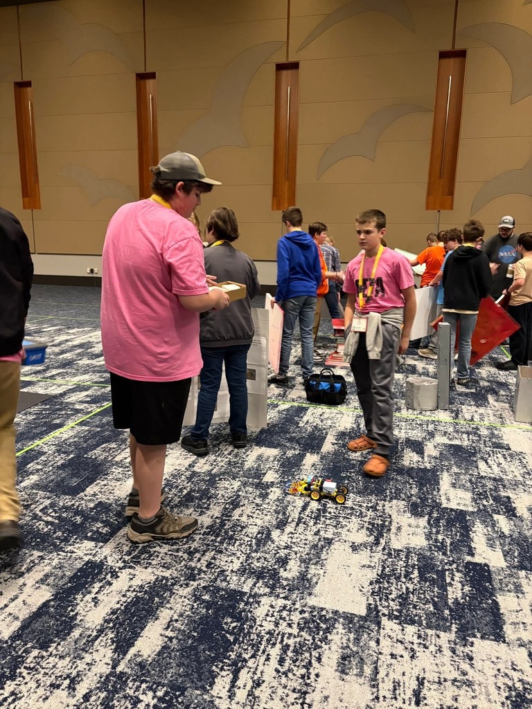 Group of people in a large room. A man in a pink shirt holds an object. Some people are wearing hats.