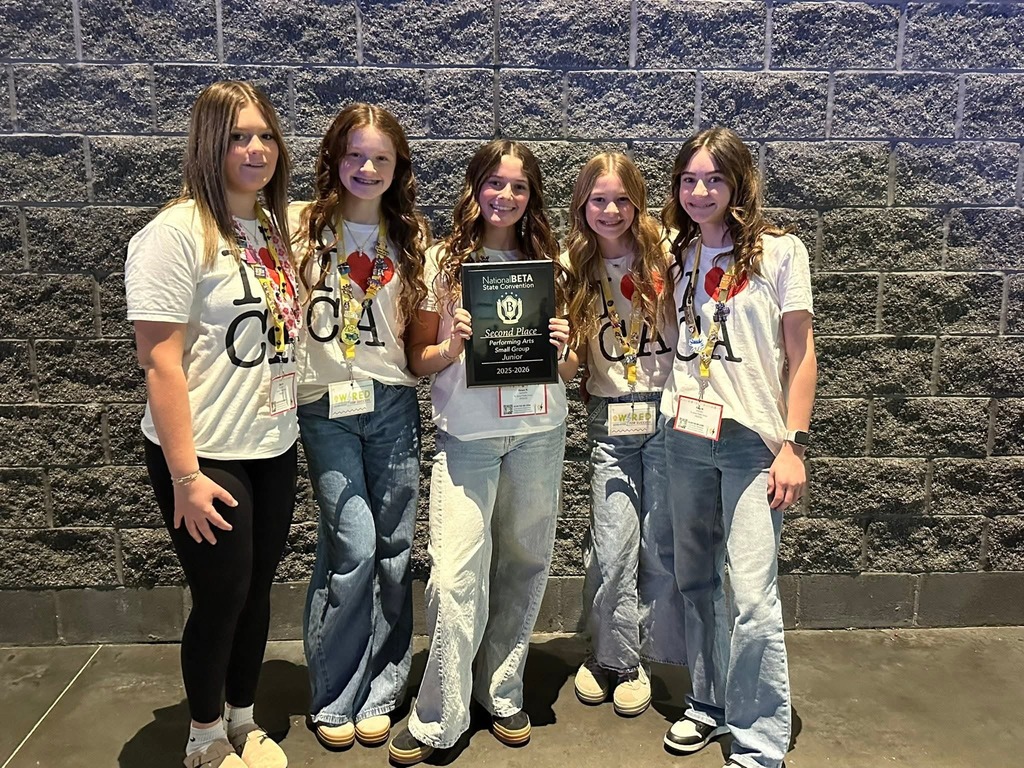 Five girls in white T-shirts and jeans stand next to each other in front of a brick wall.