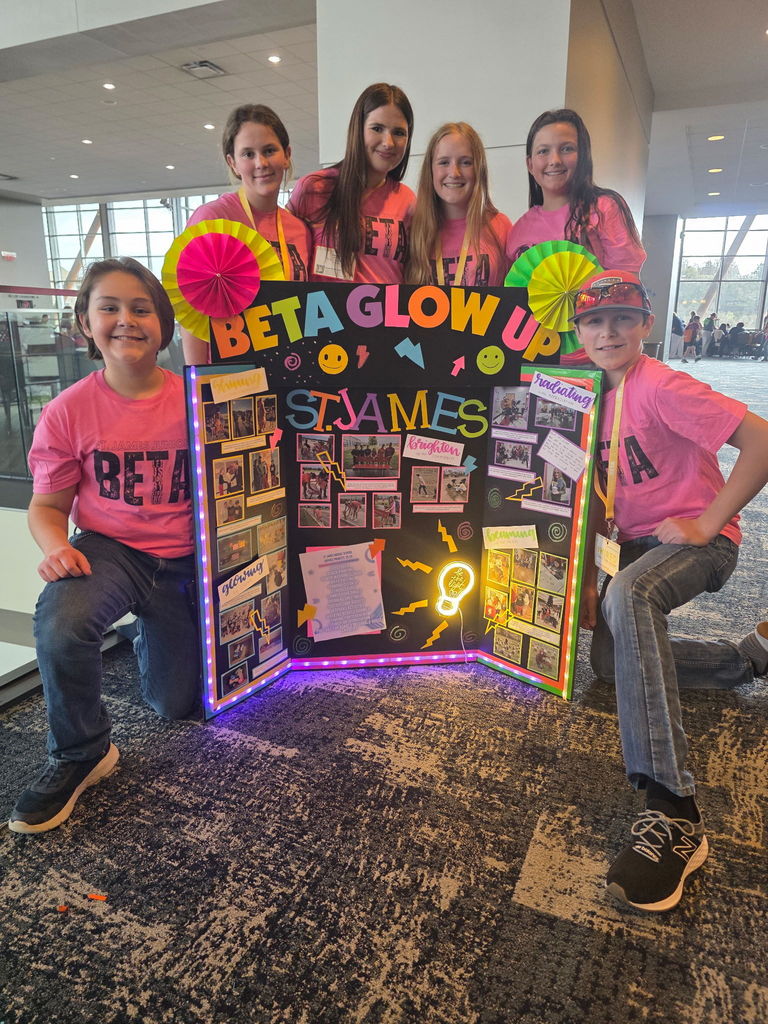 Six individuals in matching pink shirts pose behind a display, which says "Beta Glow Up". Bright colors and lights are visible.