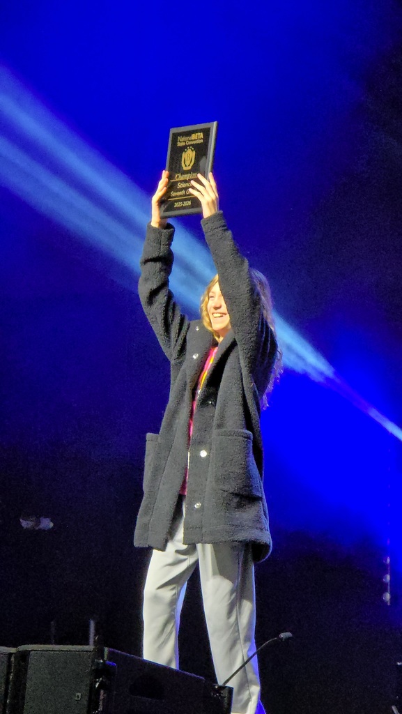 Person on stage holding an award above their head; blue stage lighting and black speaker in the foreground.