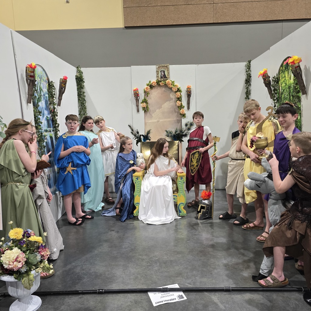 People in togas stand on a stage with one person in a white dress. Flowers and props adorn the background.