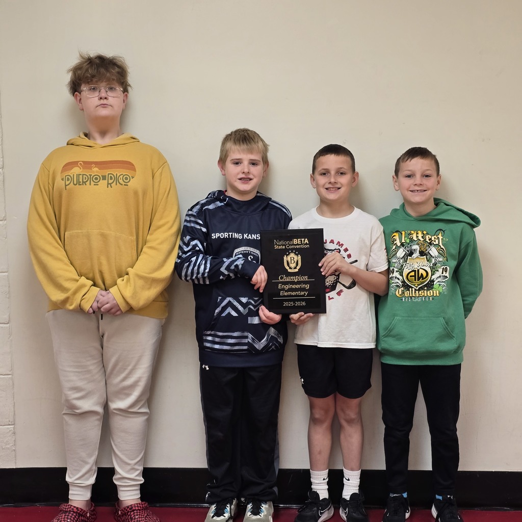 Four boys in hoodies and sneakers stand in a row, one holding a framed plaque. One wears a yellow hoodie, another a blue hoodie, one a white shirt, and the last a green hoodie.