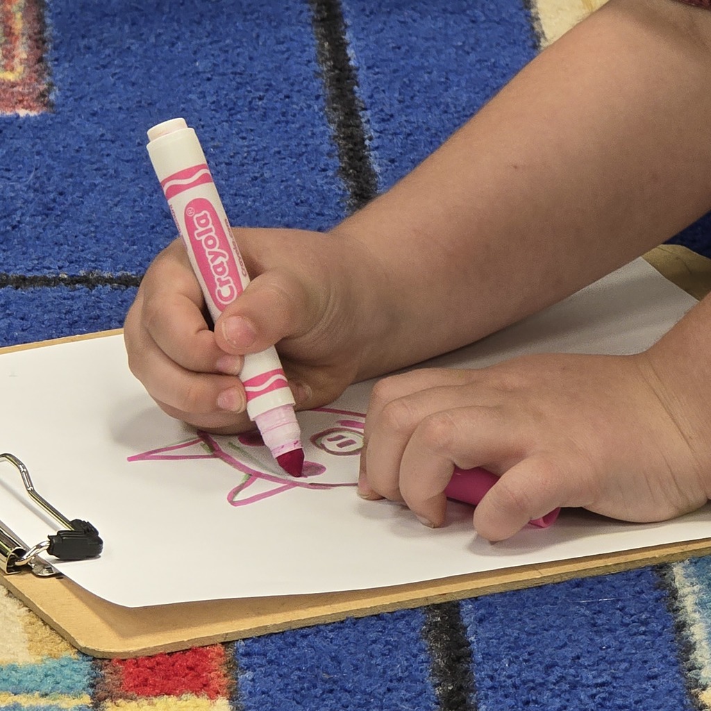 A child holds a pink marker, drawing a pink fish on a white paper on a wooden clipboard.