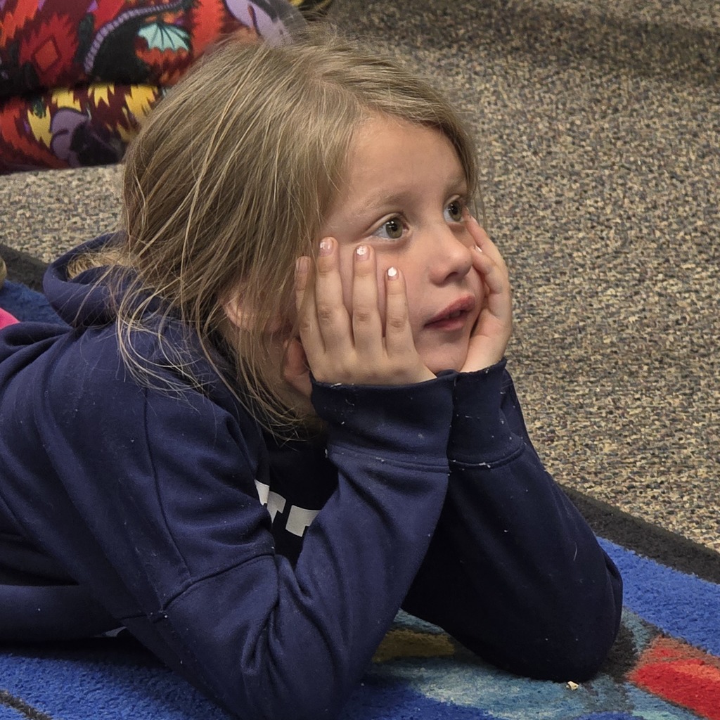 A young girl lies on a colorful rug, covering her face with her hands. She wears a dark blue hoodie.