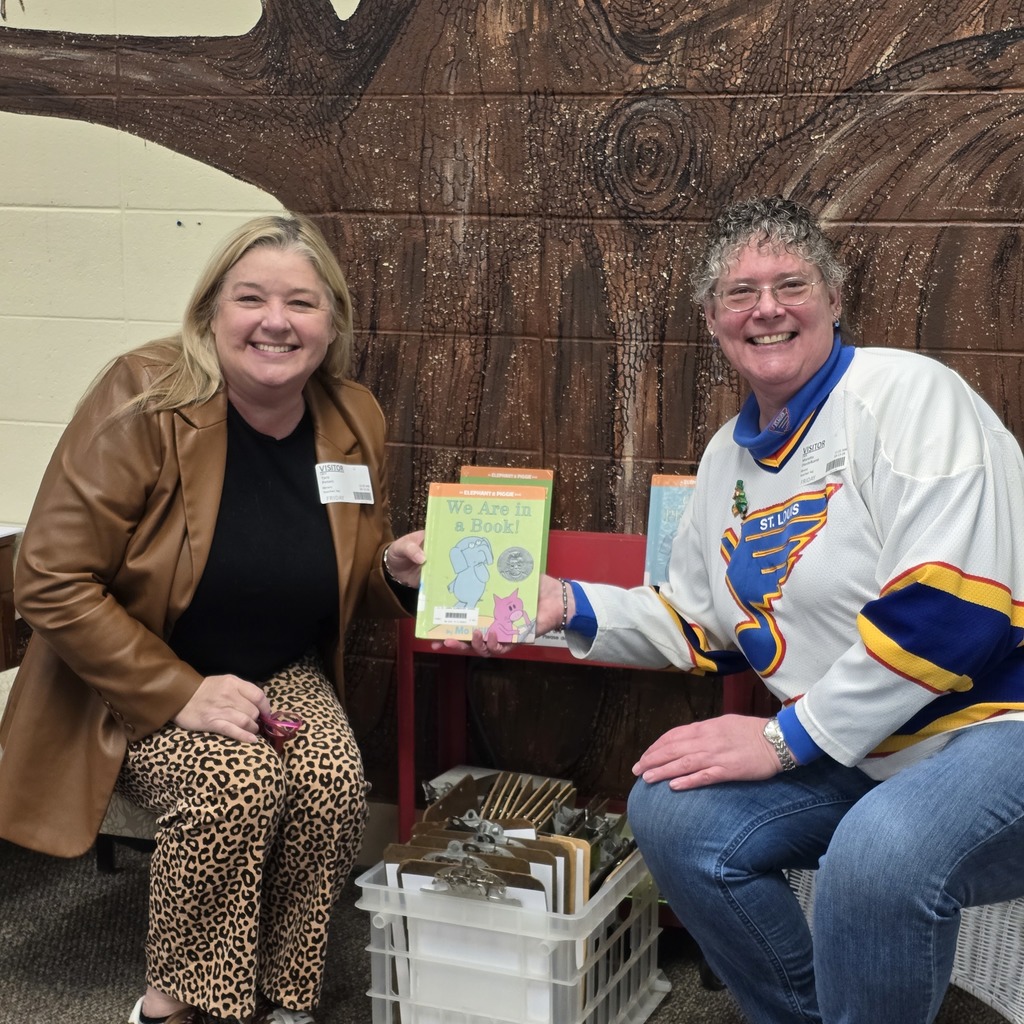 Two women seated, one holding a book, other in hockey jersey, smiling at camera. Background features mural.
