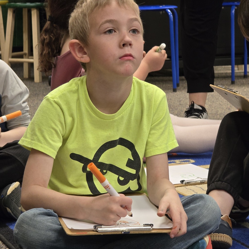 Child with short hair in a yellow shirt seated, holding a marker, drawing in a notebook.
