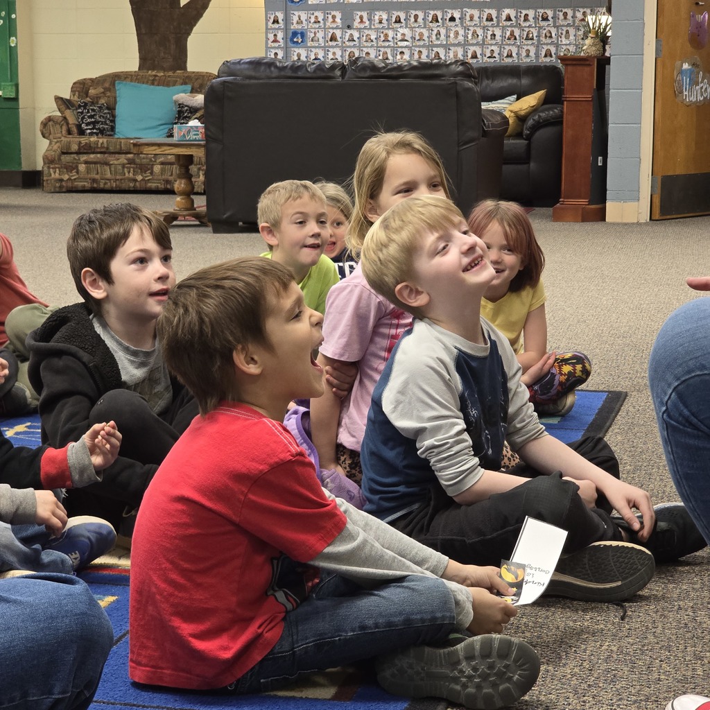 Several children sit on the floor. One holds a paper. Others are looking at a person in front.