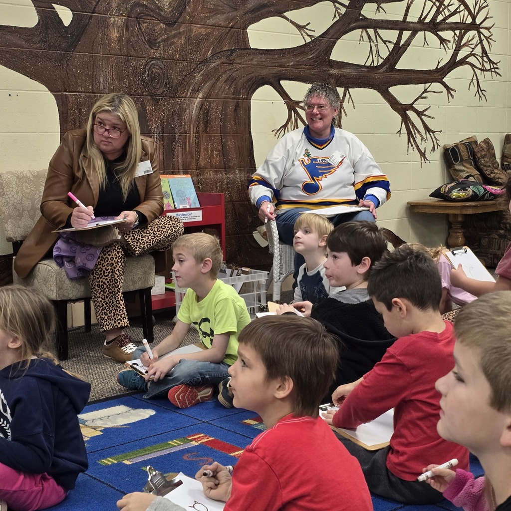 A group of children and two women sit on a rug. One woman writes on a pad.