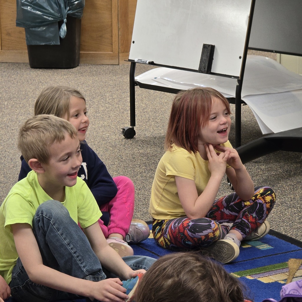 Four children sit on a rug, smiling. A wooden cabinet and trash bin in the background.