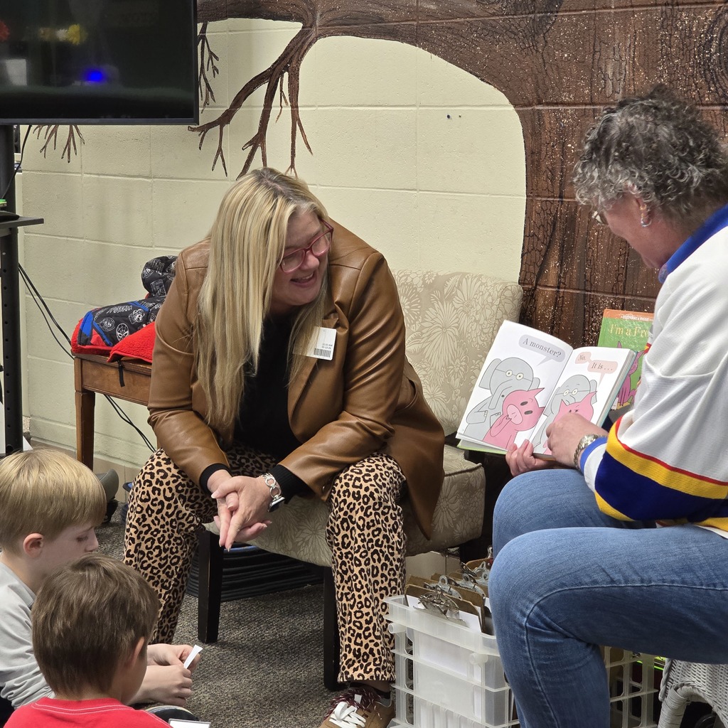 Woman sits with two children; one reads aloud from a book. Behind them, another woman smiles.