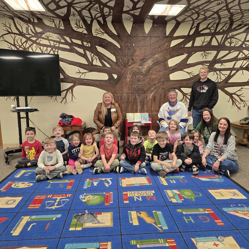 A group of children and adults are sitting and kneeling on a colorful carpet in a room with a tree mural.