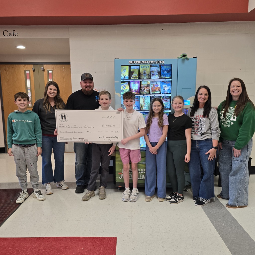 A group of adults and children pose for a photo with a check in a school hallway.