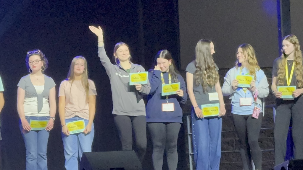 Eight people, some holding yellow items, stand on a stage with blue lighting. One person waves.