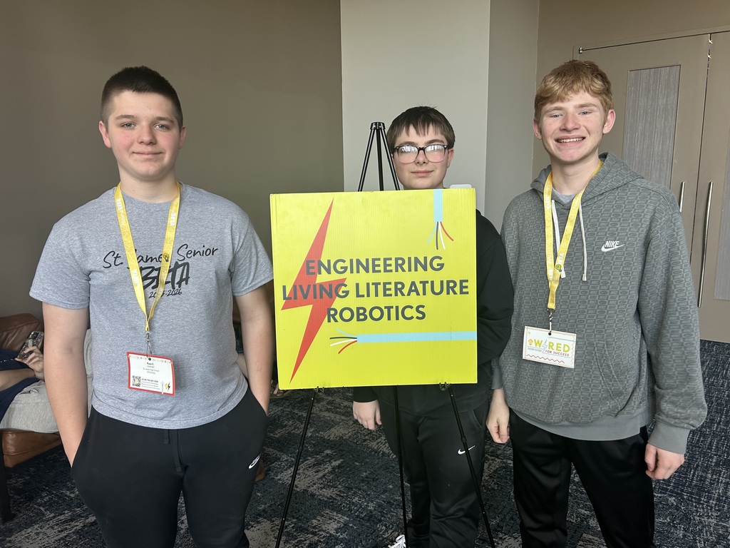 Three boys stand next to a yellow sign with a lightning bolt. Two wear gray t-shirts and one wears a gray hoodie.