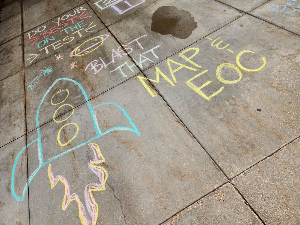 Sidewalk with various colorful chalk drawings, including a rocket and text that reads "Blast off that mat log."