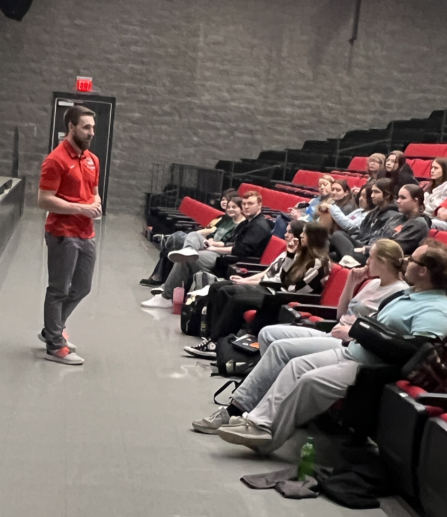 Auditorium with a man standing and speaking; seated audience on red chairs with bags and items on the floor.