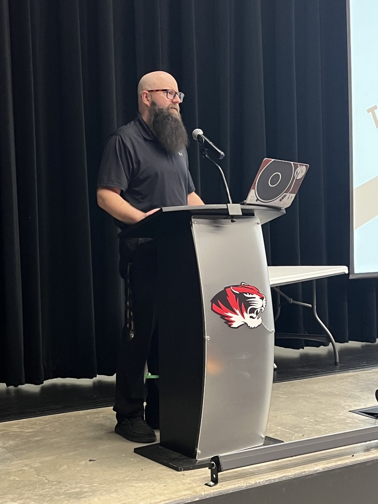 Man with a beard stands at a podium with a laptop. Microphone in front, black curtain in the background.