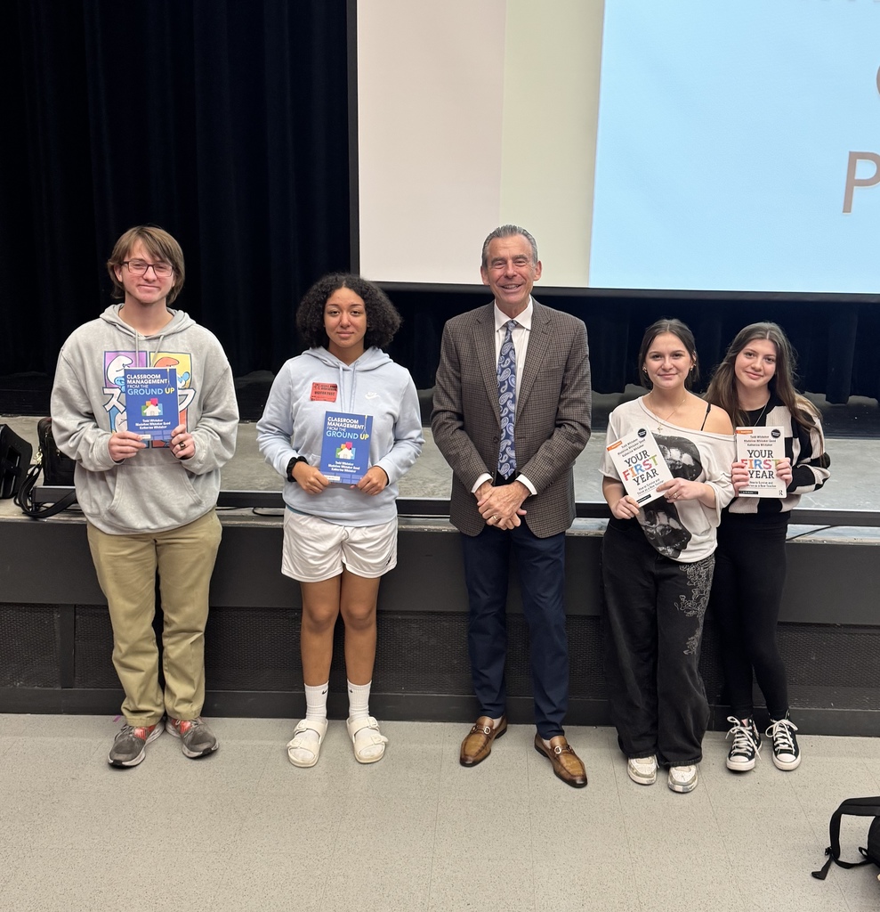 Five people, three holding books, stand on a stage with a projector screen behind them.