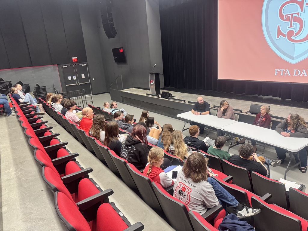 Auditorium with rows of red chairs and people seated. A screen displays "FTA DA". Some individuals at a table.