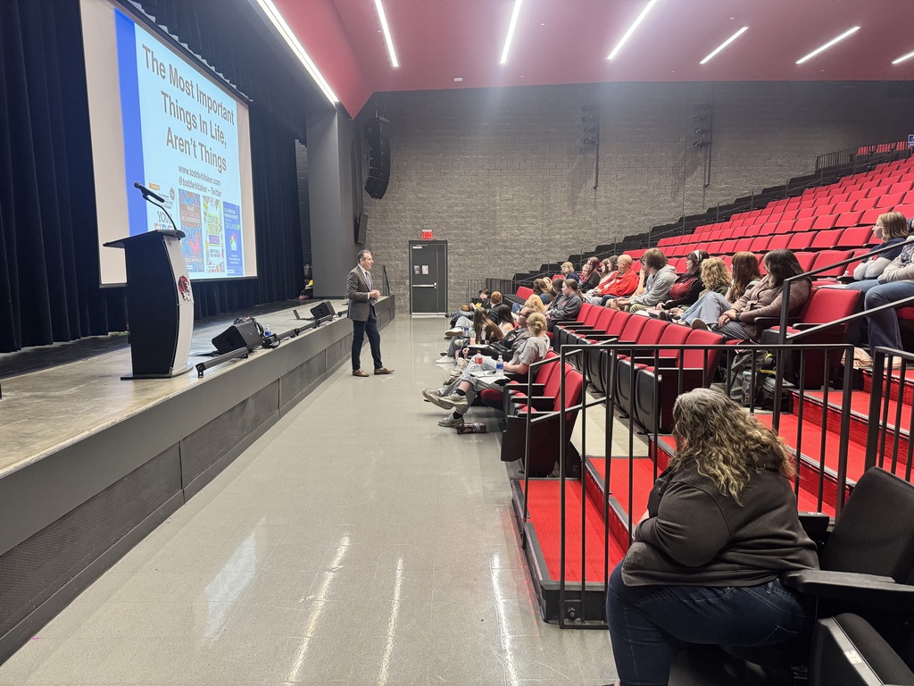 Person stands on stage with a microphone in an auditorium. An audience sits in red seats.