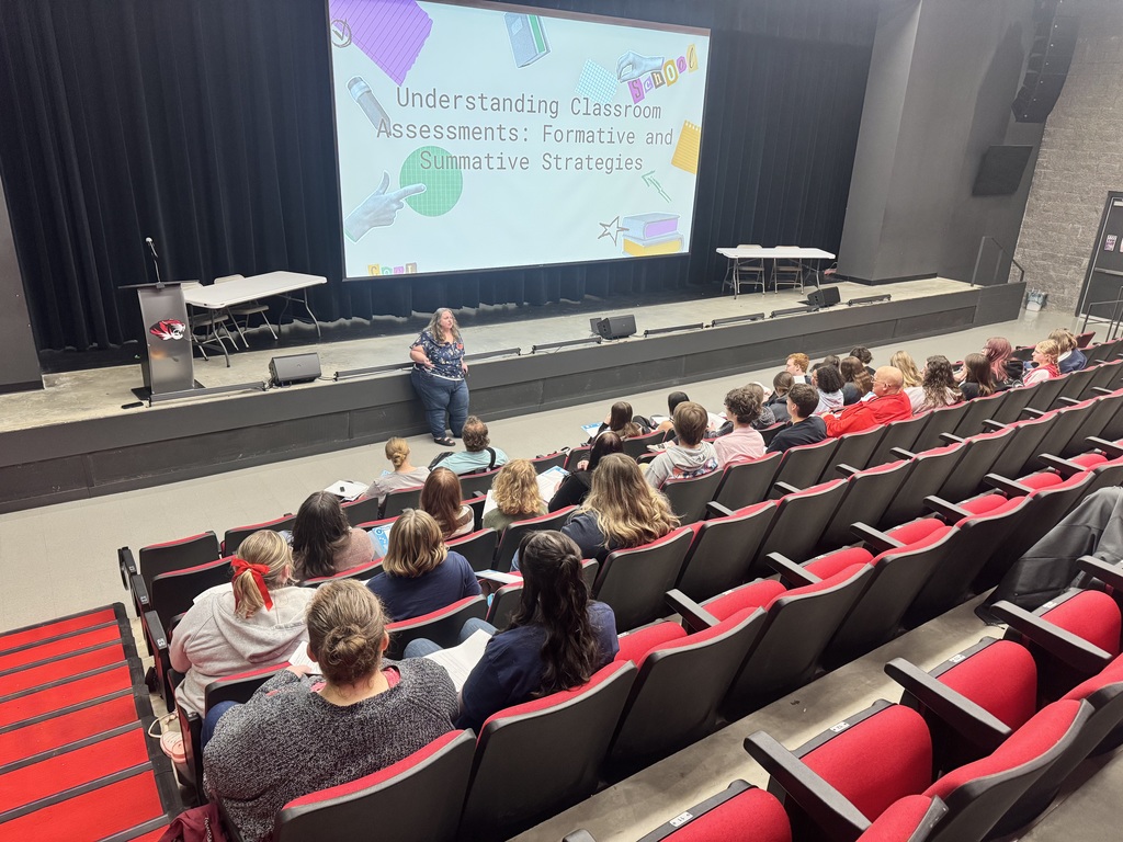 An auditorium with red seats, a person sitting in front, and a screen with a presentation.