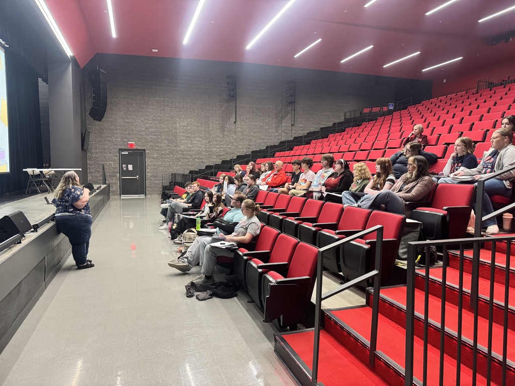 A large room with red chairs and metal railings. A person is seated on the left, and a few people are seated in rows.