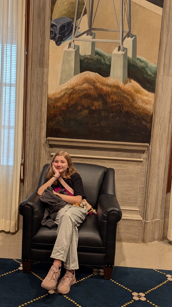A woman sits on a chair in a room with a mural of a landscape. She is smiling.