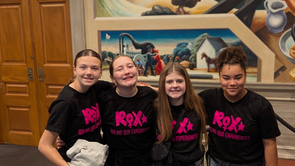 Four girls in matching black t-shirts with pink text pose for a photo in front of a mural.