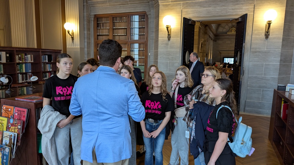 People in black t-shirts stand in a library. A man in a blue shirt addresses them. Behind them, a bookshelf and an open doorway with lit sconces.