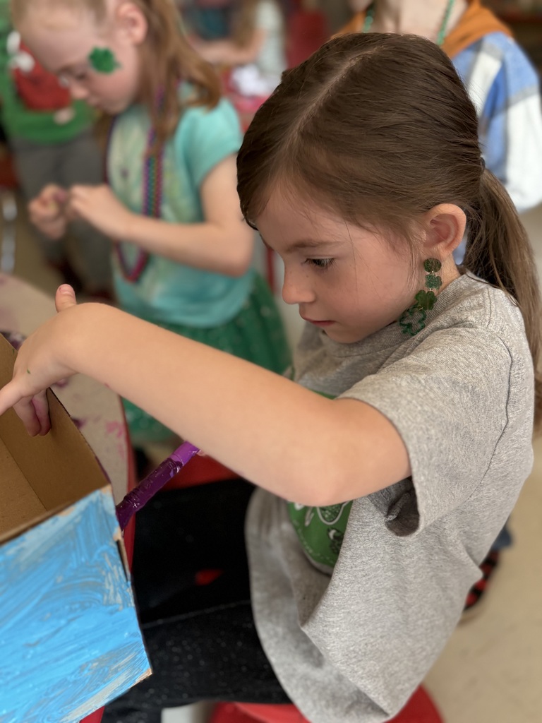 Girl with ponytail seated on chair, pointing to blue painted box while another child stands behind.