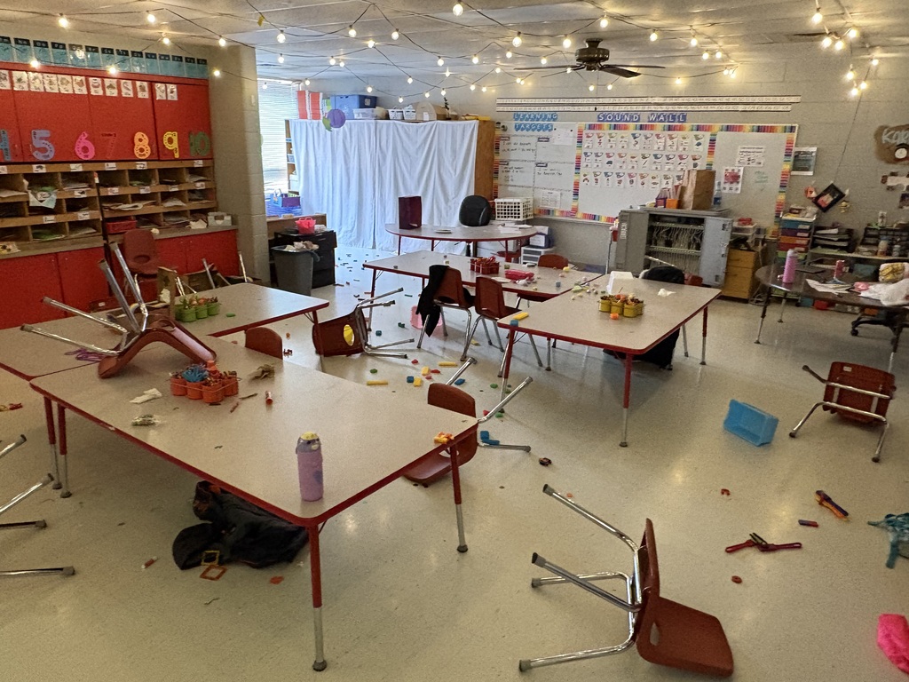 A classroom with overturned desks, chairs scattered, and a messy floor, with a white backdrop.