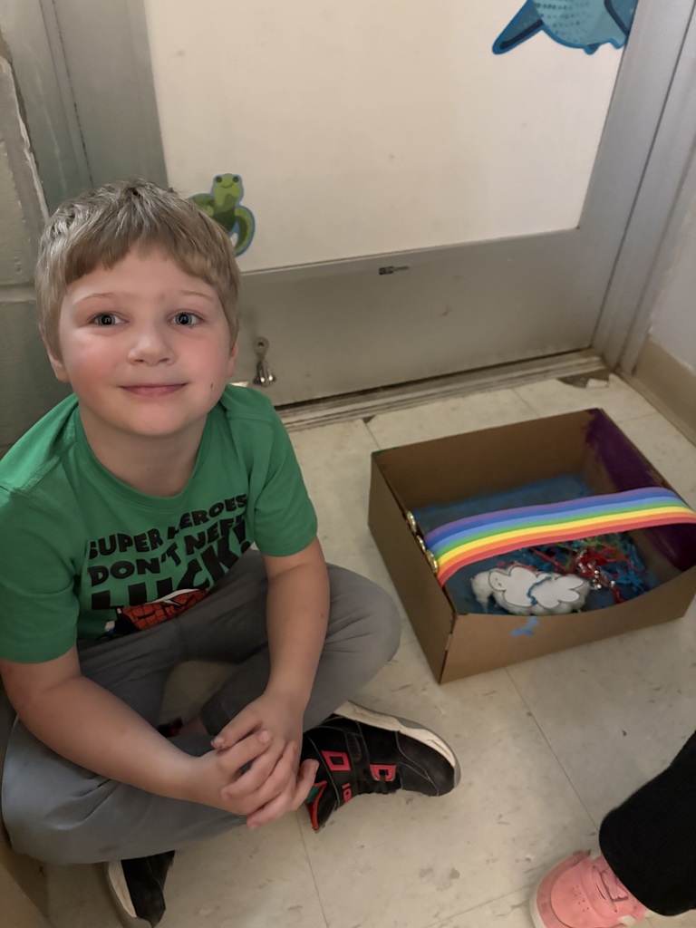 A boy wearing a green shirt sits on the floor next to a box filled with toys.