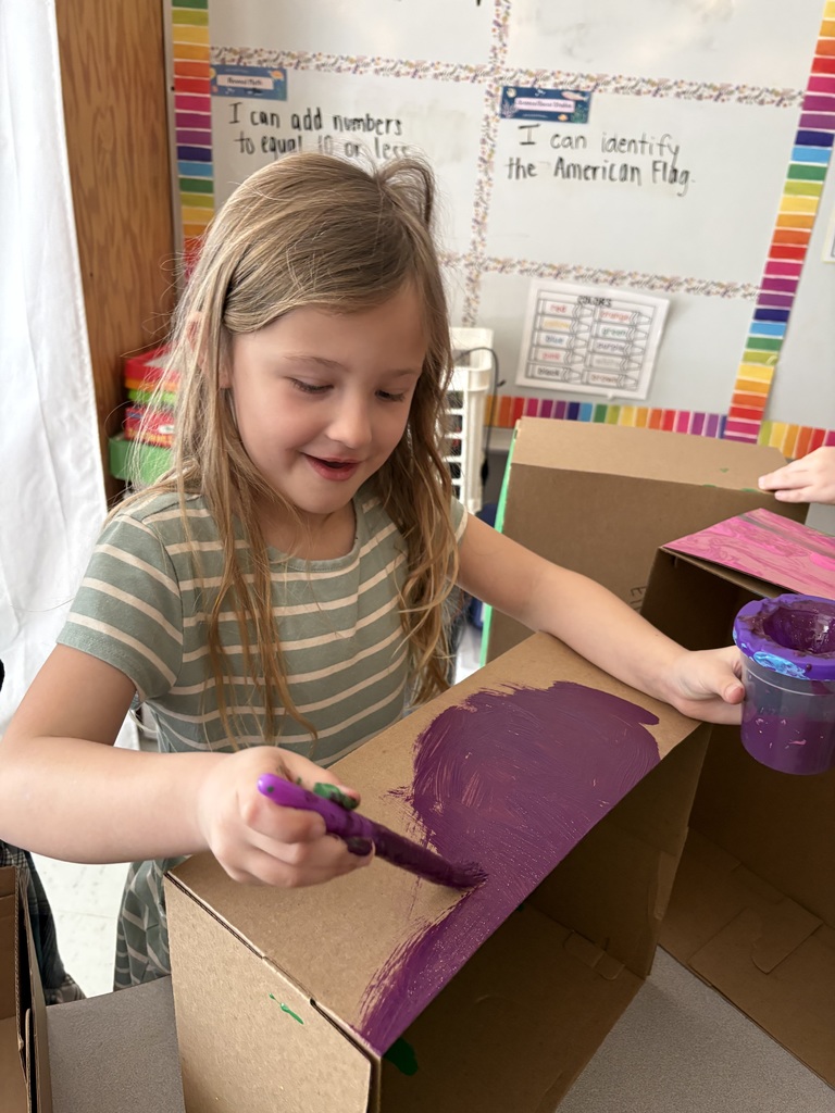 A girl uses a paintbrush to apply purple paint to a cardboard box, standing in a classroom with a bulletin board and shelves in the background.