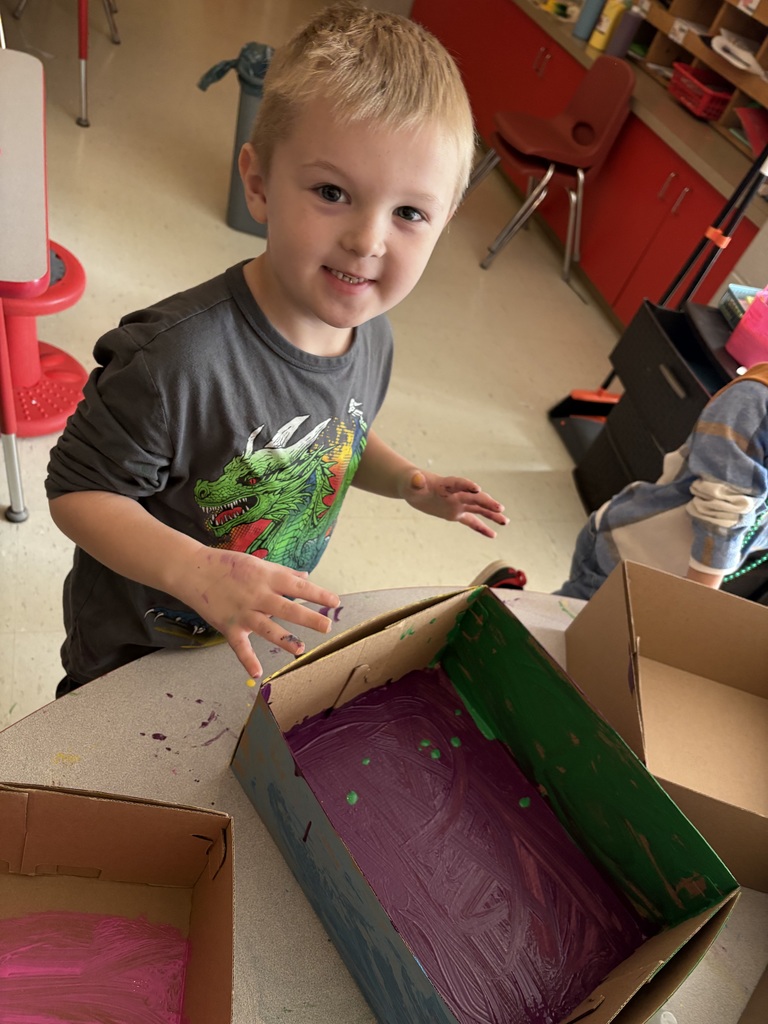A child with blond hair is standing at a table with hands covered in paint. Two cardboard boxes are on the table.