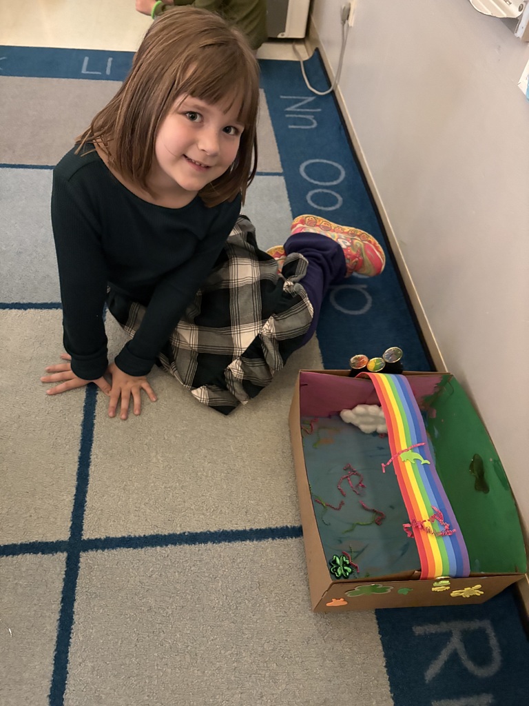 A young girl sits on the floor next to a box with a rainbow. She is wearing a black shirt and a plaid skirt.