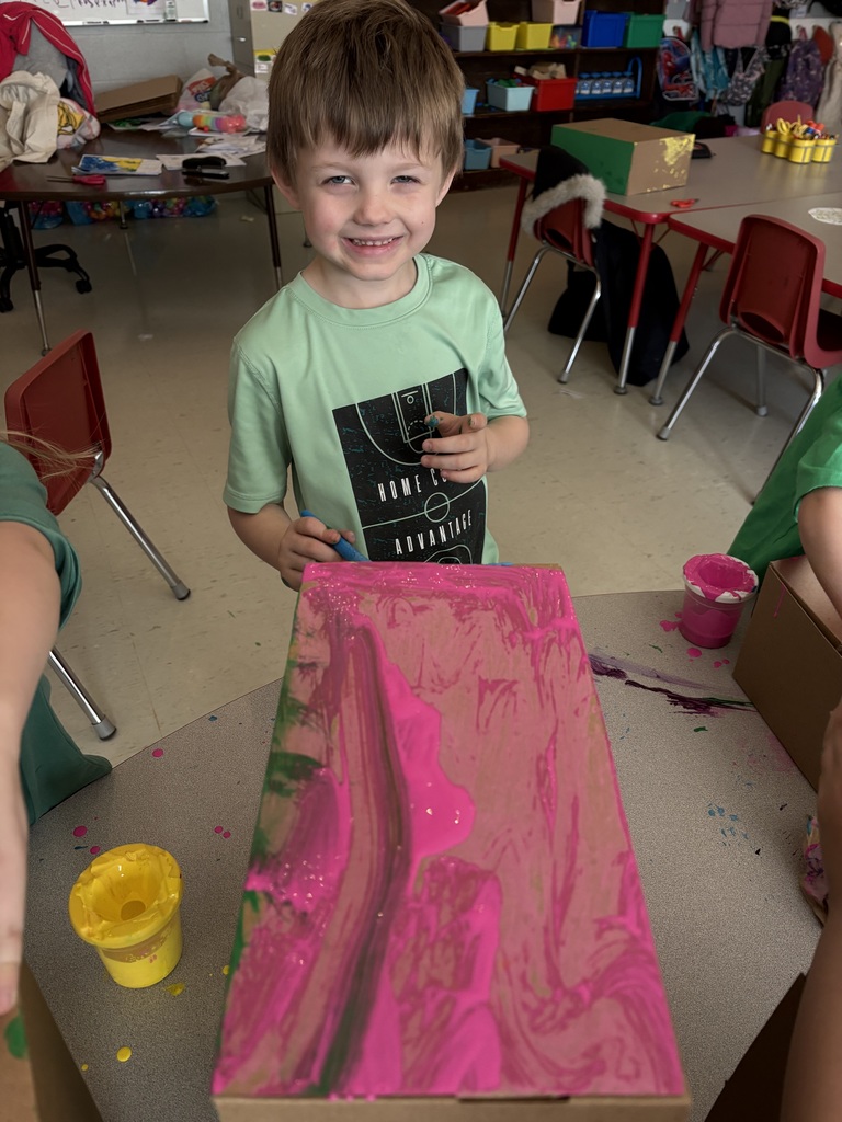 A child stands with a pink marbling project, wearing a green t-shirt, in a classroom setting.