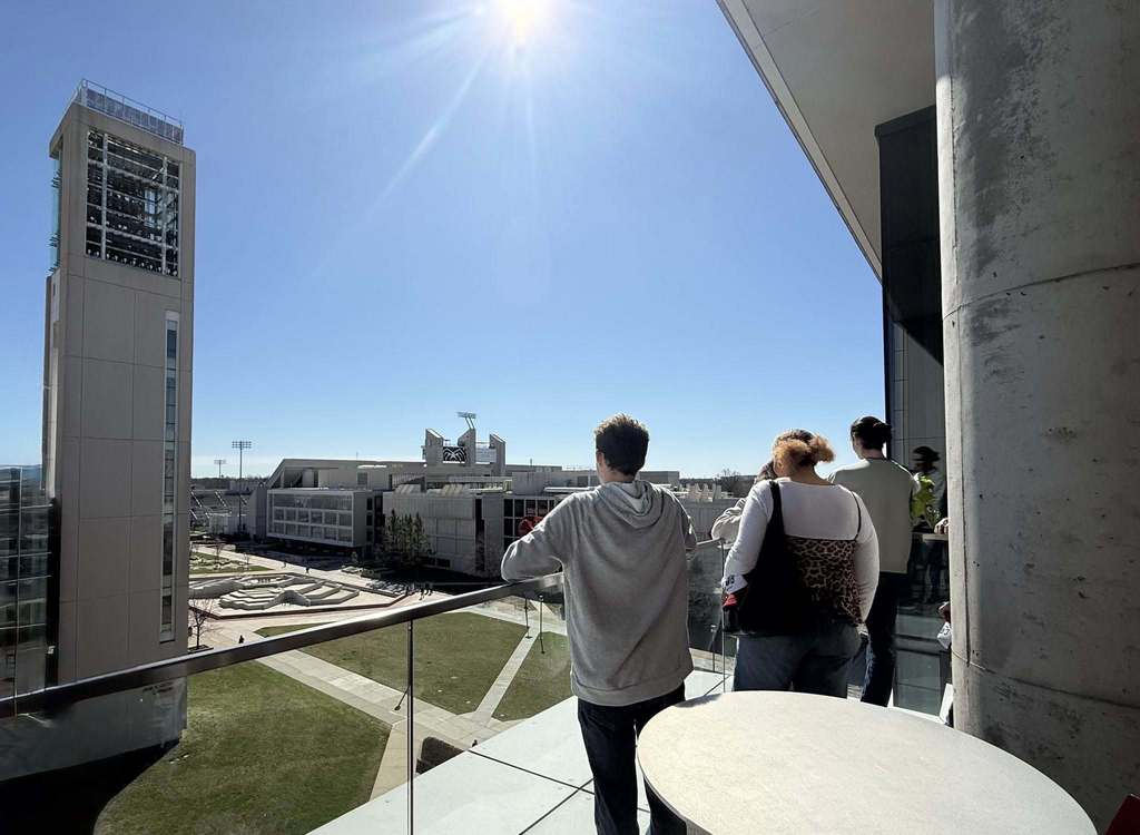 People stand on a balcony with a grassy lawn below, a tower behind, and city buildings in the distance under a bright sky.
