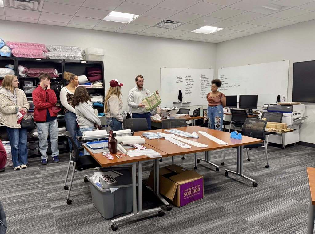 Nine people stand around a table in a classroom with whiteboards, a monitor, and shelves of materials.