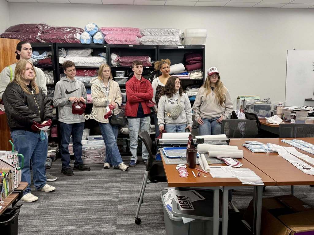 A group of people in a room with shelves and desks, some holding items, others listening.