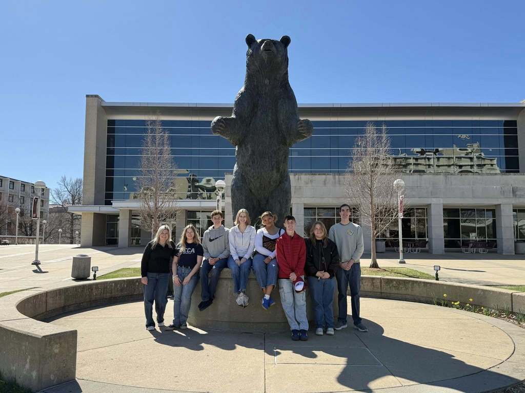 A group of people stands around a large bear statue in front of a large building.