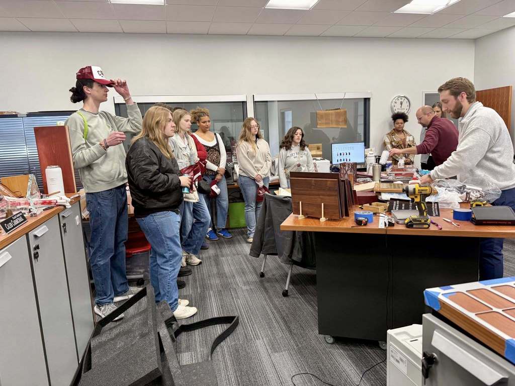 Office with white walls, gray floors. People gather around a desk, some using equipment, others watching. Desk holds monitors, bottles, and other objects.