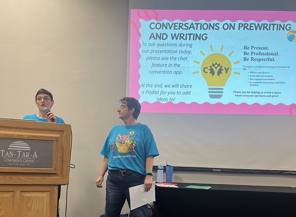 Two people in blue T-shirts stand at a podium in front of a projector screen displaying "Conversations on Prewriting and Writing".