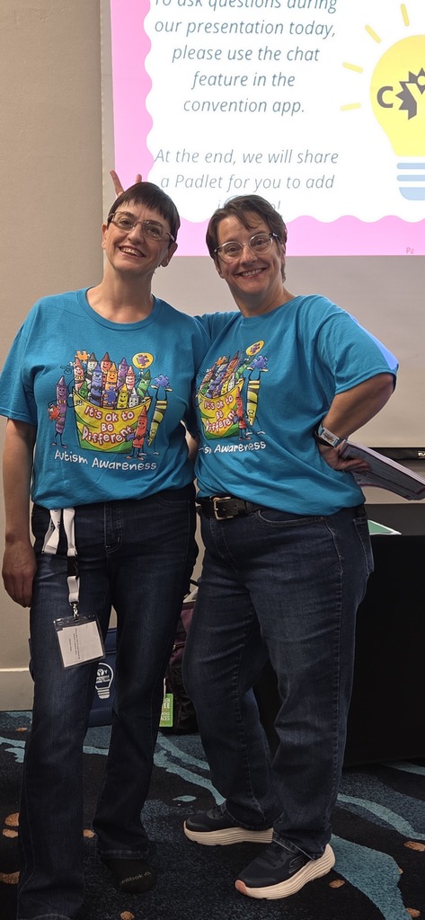 Two women wearing blue t-shirts stand next to each other, smiling. They have name tags and are in front of a screen with text.
