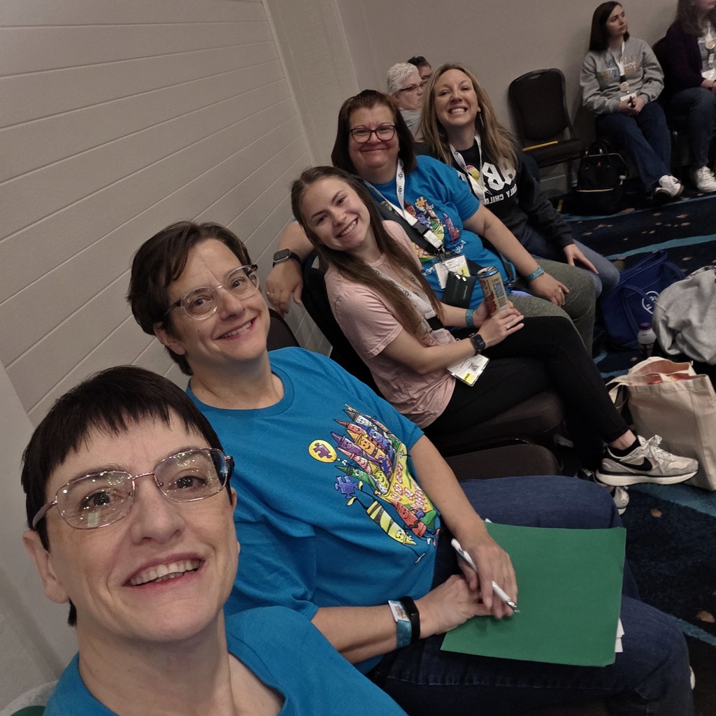 A group of smiling adults in blue shirts and glasses sit in chairs, one holding a pen and a folder.