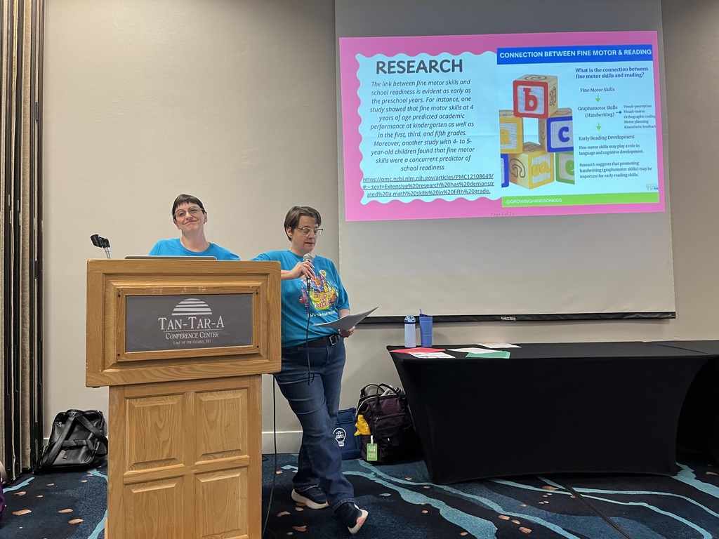 Two women wearing blue t-shirts stand next to each other, smiling. They have name tags and are in front of a screen with text.