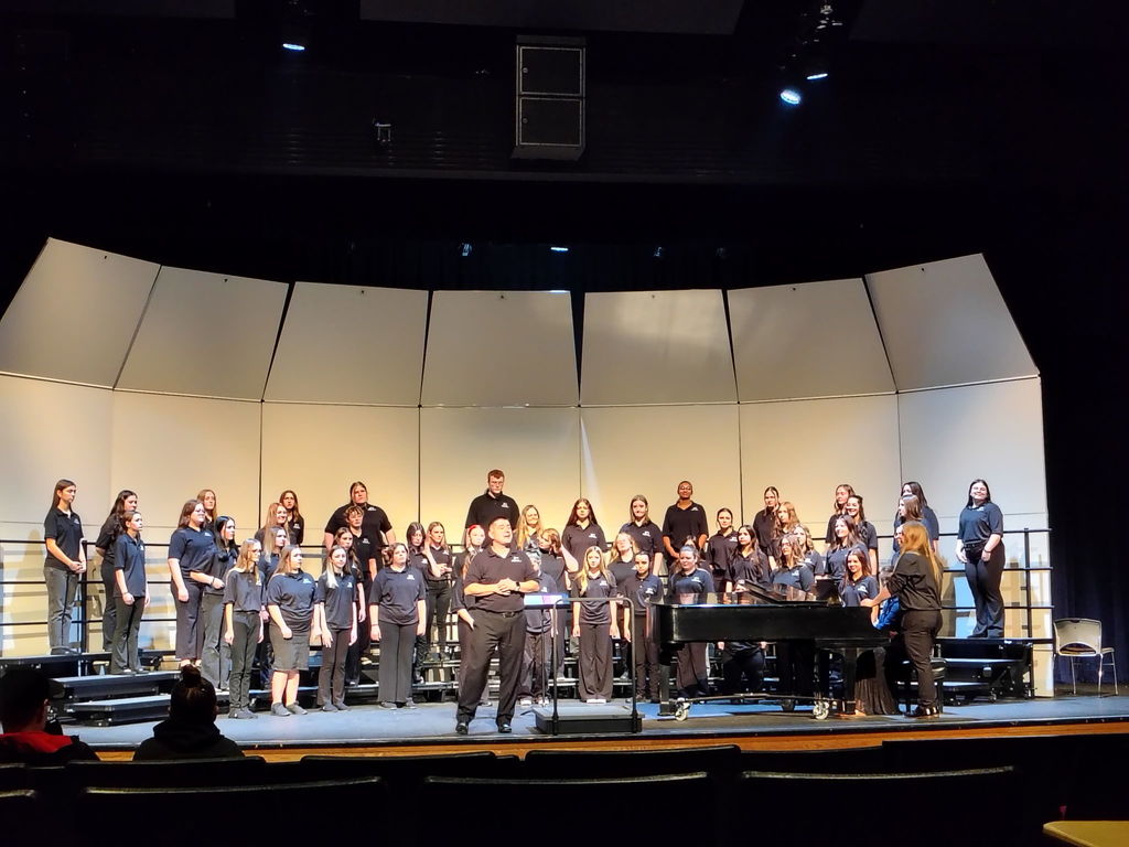 A group of people in matching uniforms stand on a stage. A piano is in the center, with others facing the audience.