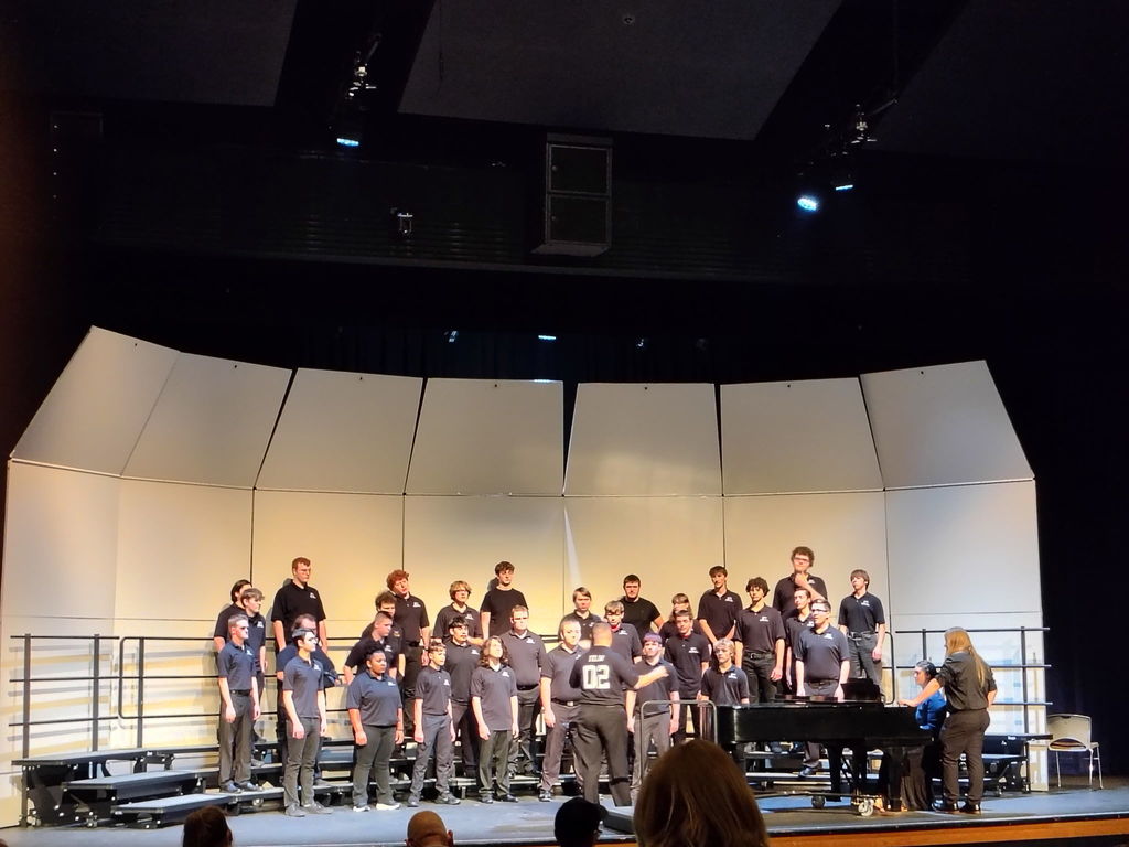 A choir stands on stage with a piano, all wearing matching shirts. Behind them, a white curved wall and a dark background are visible.